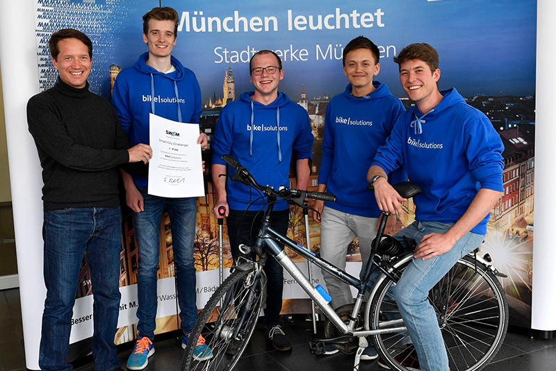 Preisträger tragen blaue Hoodies und stehen hinter einem Fahrrad, neben ihnen ein Vertreter der Stadtwerke München