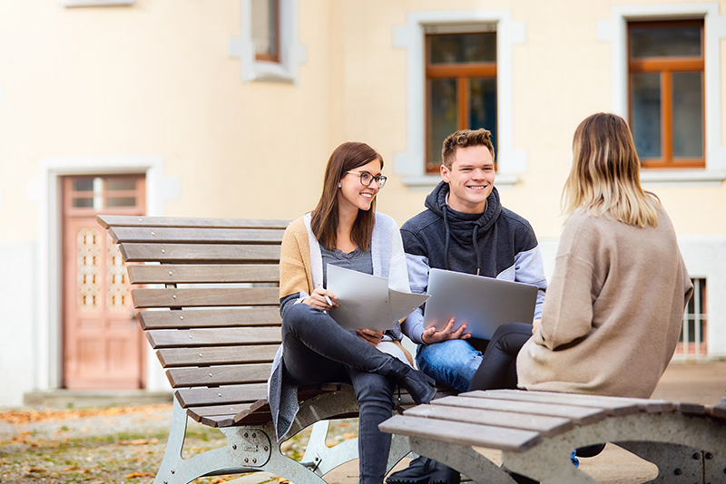 Zwei Studentinnen und ein Student sitzen auf Holzbänken