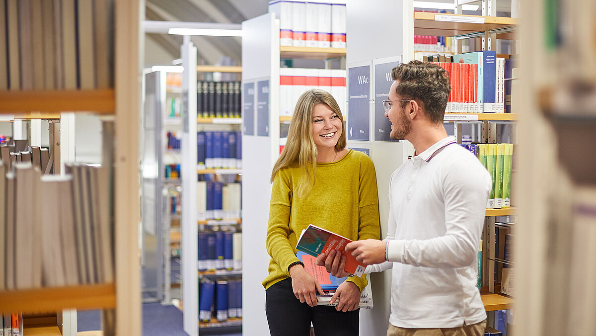 Zwei Studierende stehen zwischen Bücherregalen in der Bibliothek und unterhalten sich. 