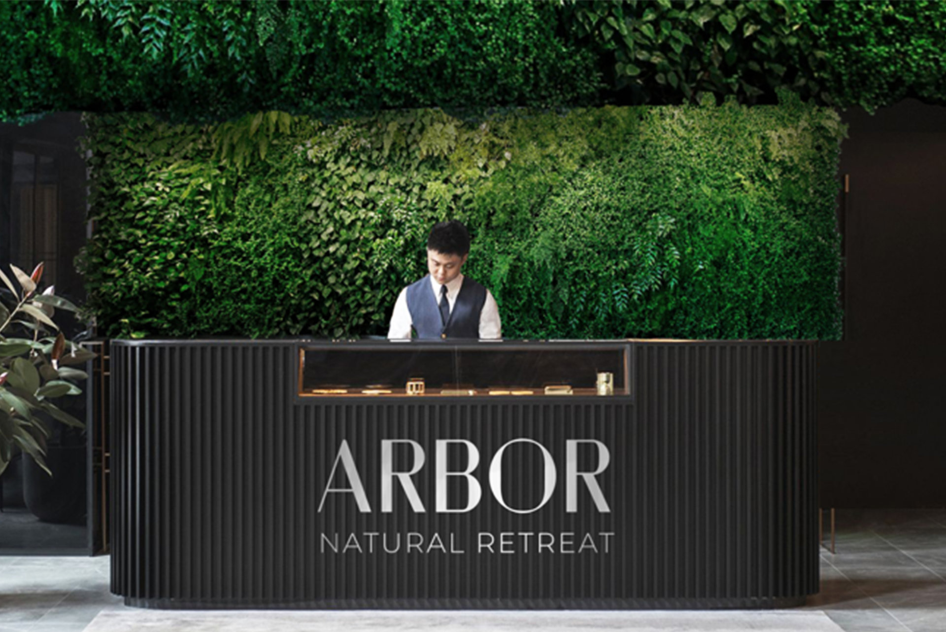 Rezeptionstresen des Arbor Natural Retreat mit Rezeptionist vor einer grünen Hecke im Eingangsbereich eines Hotels