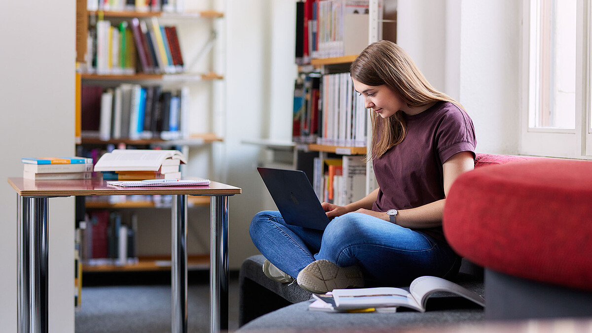 Eine junge Frau sitzt in der Bibliothek und arbeitet an ihrem Laptop.
